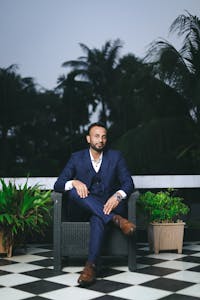 Man in formal attire seated outdoors on a patterned floor with plants.